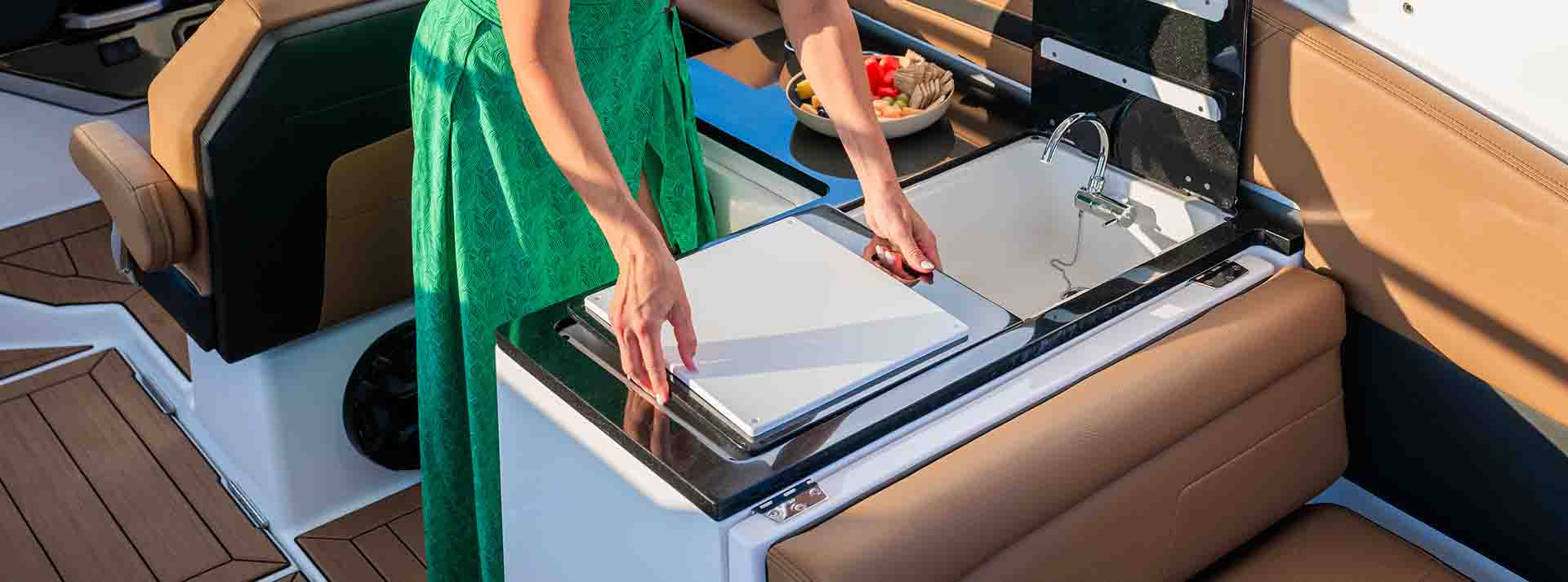 Woman preparing food at the integrated galley station aboard the Yamaha 295 Series, featuring a sink and prep surface next to plush brown seating.