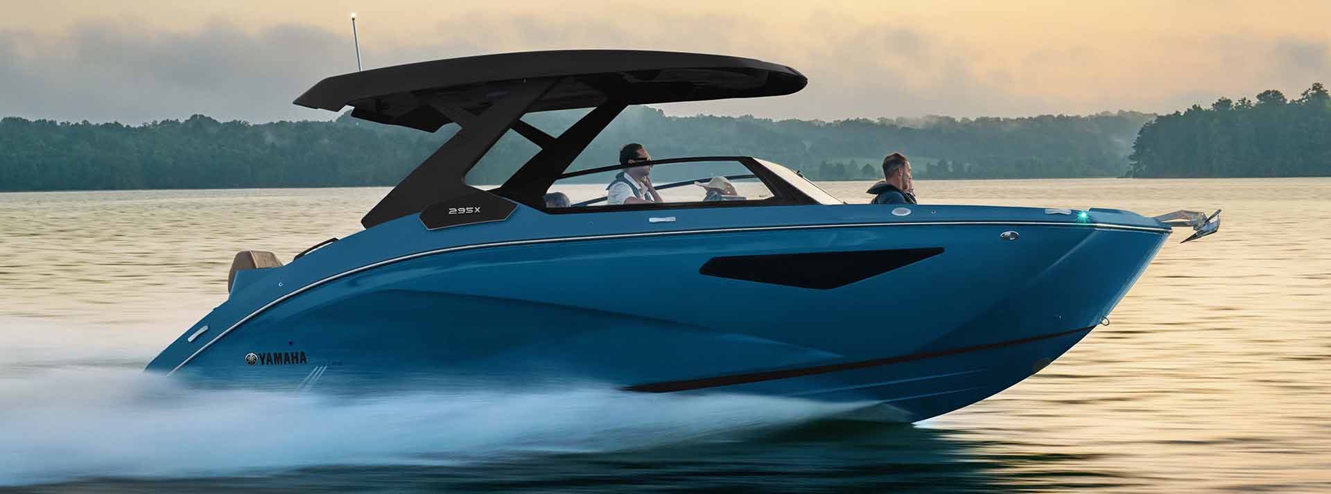 2025 Yamaha 295X cutting through the water at high speed, showcasing its sleek blue hull design and black hardtop against a scenic lake backdrop at sunset.