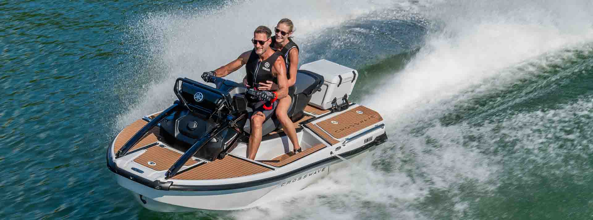 A couple riding a Yamaha Crosswave at high speed on open water, with water spraying dramatically behind them. Both riders are wearing Yamaha-branded life vests.
