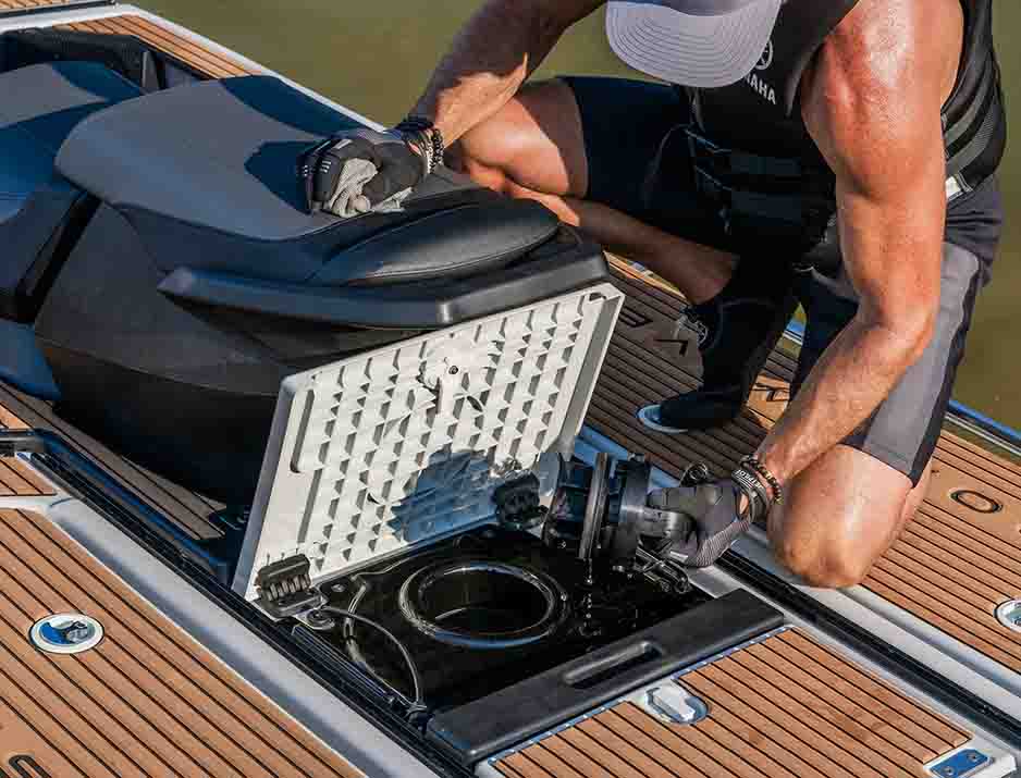 Man kneeling on the deck of a Yamaha Crosswave while opening a rear maintenance hatch, exposing internal mechanical components.